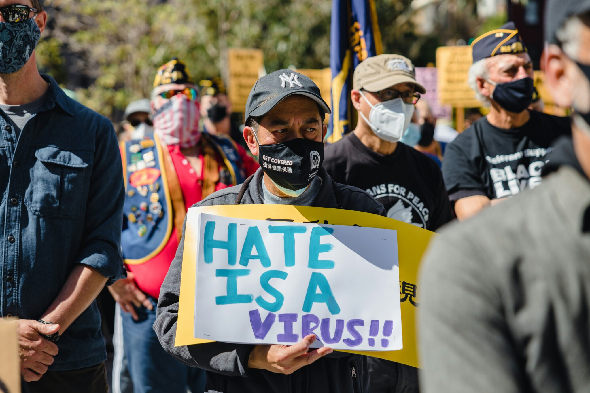 man in a mask holding a sign that says "HATE IS A VIRUS"