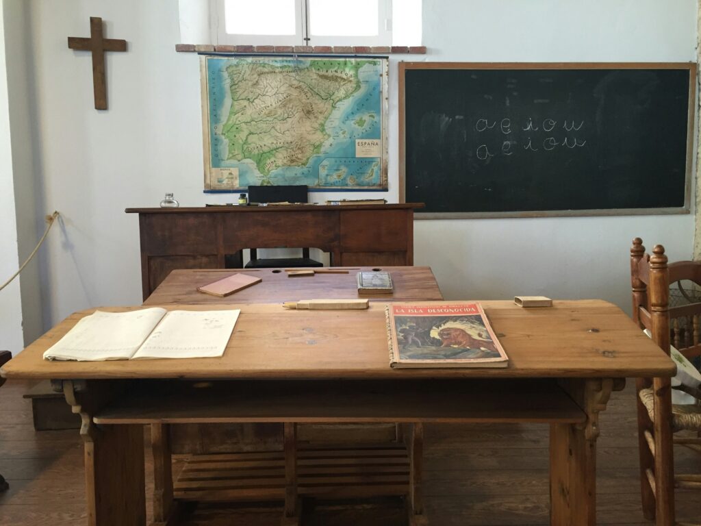 A cross in a classroom with a map, a blackboard, and a desk