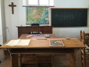 A cross in a classroom with a map, a blackboard, and a desk